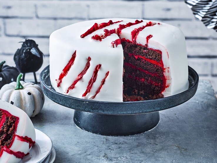 Auf einem Tortenteller steht eine gruselige Halloween-Torte. Sie ist mit weißem Fondant eingedeckt und hat blutige Risse als Dekoration.