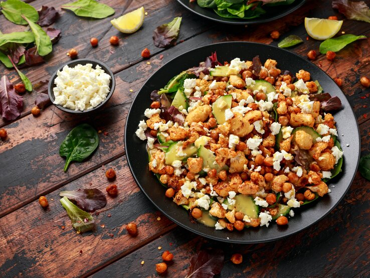 Eine große schwarze Schüssel mit dem gerösteten Blumenkohl-Kichererbsen-Salat mit Gurke und Feta neben einer kleinen Schüssel mit zerbröseltem Feta und vielen losen Salatblättern und Zitronenspalten, vor einem Holzhintergrund von schräg oben. 