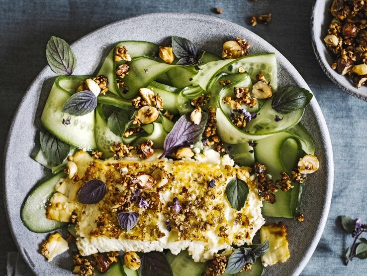 Gratinierter Feta mit Ahornsirup-Nusscrunch von oben mit Gurkengemüse in einem hellgrauen Teller neben einer Schüssel mit Nüssen auf einem grauen Tisch. 