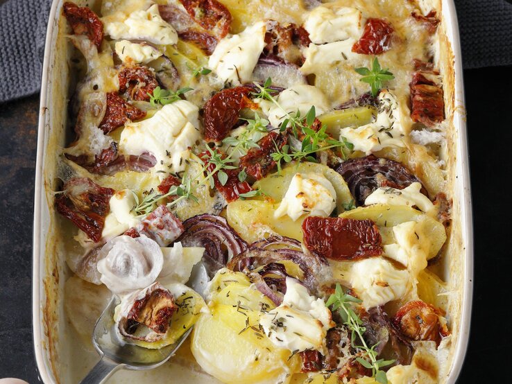 Kartoffelgratin mit getrockneten Tomaten, roter Zwiebel und Thymian, mit Käse überbacken in einer großen Auflaufform auf einem dunklen Tisch. 