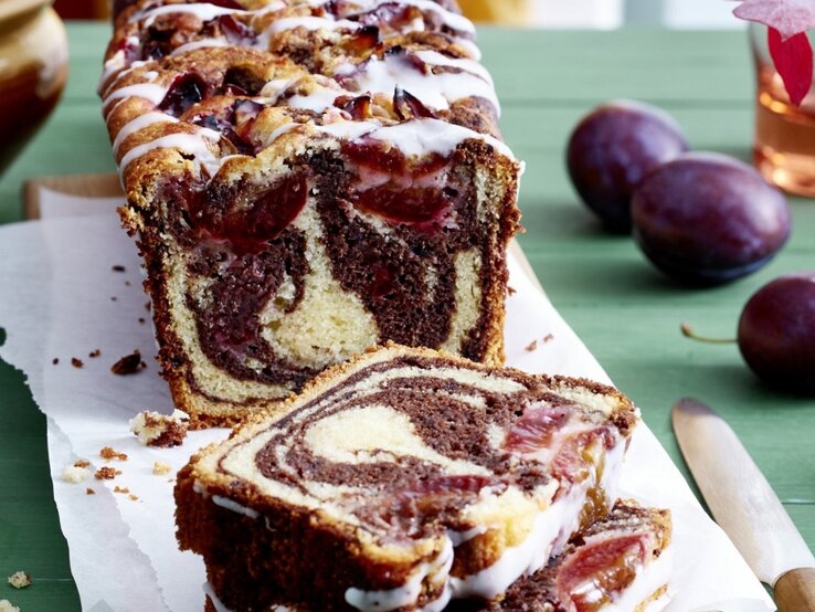 Auf einem Schneidebrett steht ein Zwetschgen Marmorkuchen. Er ist mit Puderzuckerguss überzogen. 