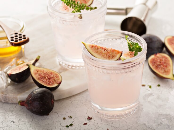 Zwei Gläser Feigen-Spritz mit frischen Feigen und Kräutern, drumherum Bar-Zubehör und Zutaten.