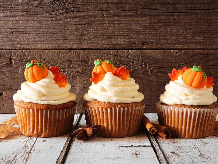 Drei Kürbis-Cupcakes verziert mit kleinen Kürbissen und Ahornblättern aus Marzipan, daneben Zimtstangen und Anissterne.