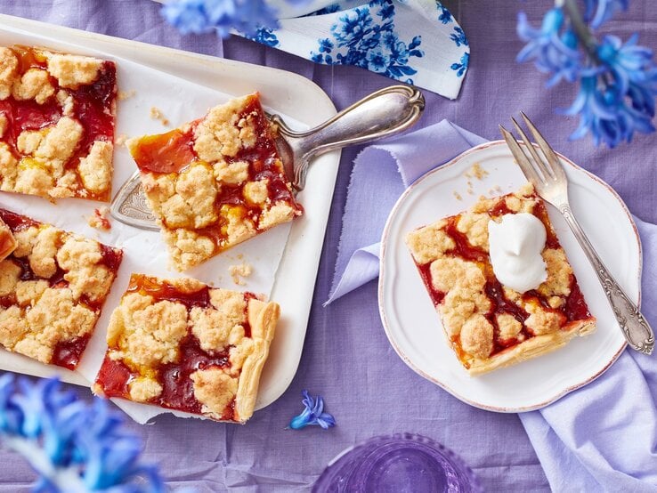 Ein weißes Kuchenblech mit Stücken des Prasselkuchens mit Erbeeren und Mango darauf neben einem Teller mit einem weiterem Stück, einer Gabel und blauen Blumen, vor einem fliederfarbenen Hintergrund, von oben. 