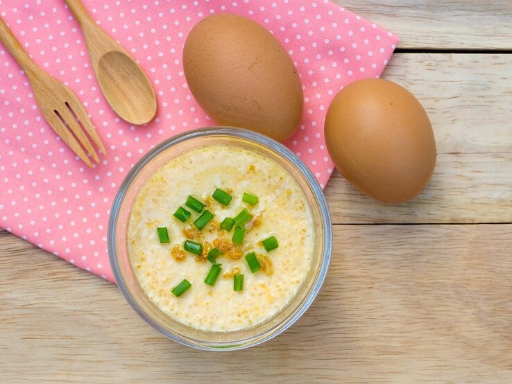 Eine Glasschüssel mit gedämpften Eierpudding mit frischen Frühlingszwiebeln auf einem roten Küchentuch neben Holzbesteck und Eiern von oben.