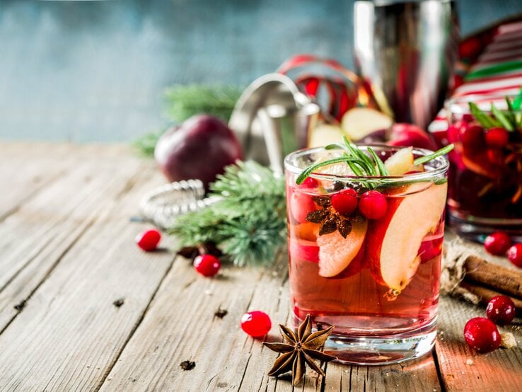 Ein Glas winterliche Sangria mit Apfel und Orange sowie Cranberrys, Anis und Rosmarin. Im Hintergrund sind unscharf die Zutaten zu erkennen.