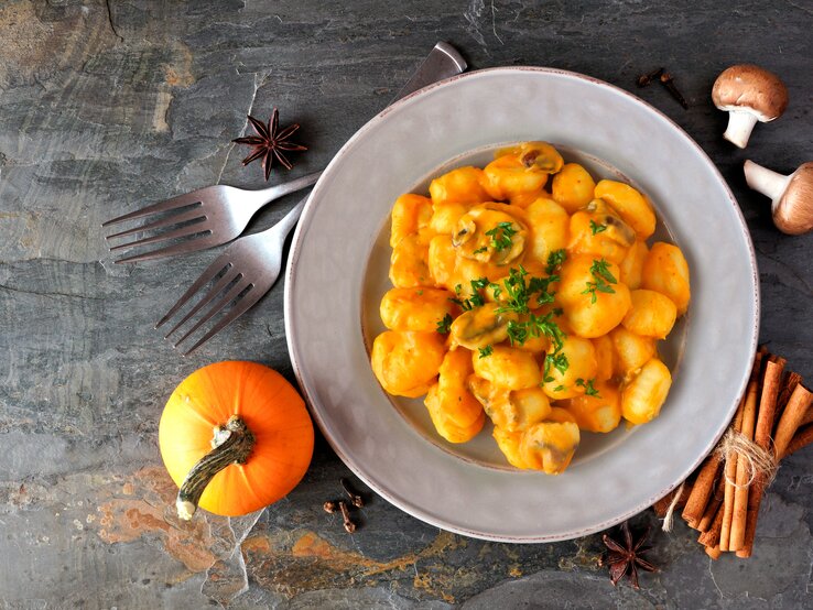 Eine Portion Kürbis-Gnocchi mit Kürbis-Pilz-Soße in einem weißen Teller steht auf einem Untergrund aus Stein. Um den Teller herum liegen Zimt, Sternanis, ein Kürbis, Pilze und zwei Gabeln.