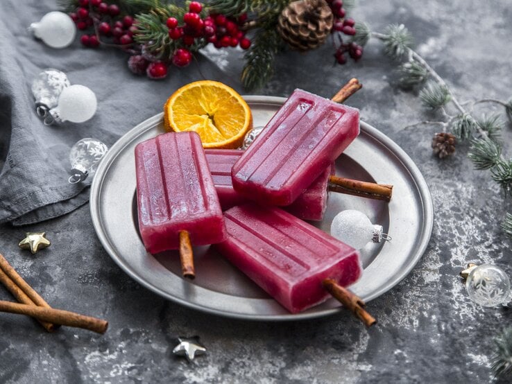 Glühwein-Eis am Stiel angerichtet auf einem silbernem Teller. Drumherum finden sich verschiedene, weihnachtliche Deko-Elemente.