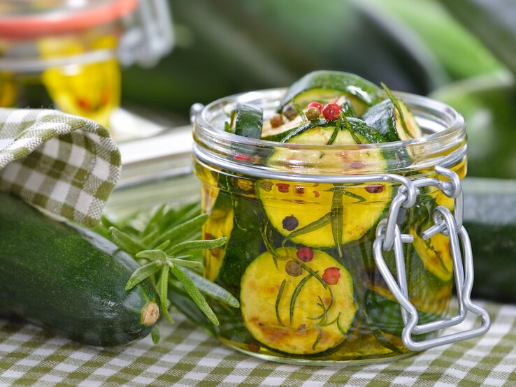 Zwei Weckgläser mit eingelegter Zucchini auf einer weiß-grün karierten Tischdecke mit Kräutern.