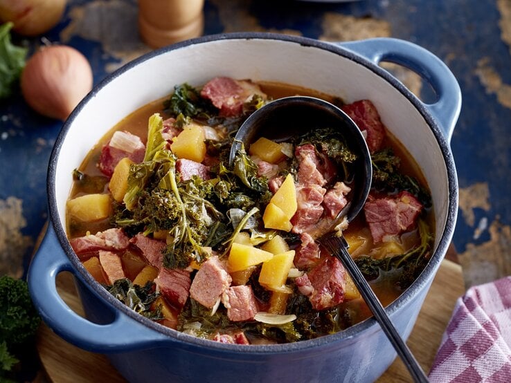 Kasseler-Gulasch in einem großen Topf mit Grünkohl, Kartoffeln und gewürfeltem Kasseler auf einer Tischplatte mit den einzelnen Zutaten drum herum. 