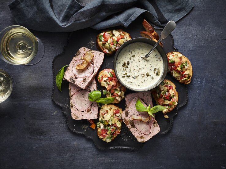 Leberkäse de luxe mit Laugen-Crostini und Artischocken-Salsa auf einer Snackplatte. Daneben zwei Gläser Sekt und ein dunkles Küchentuch. 