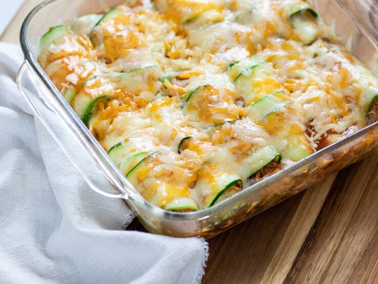 Überbackene Zucchini-Enchiladas mit Hack in einer Auflaufform. Daneben ein Tuch. Das Ganze auf einem hölzernen Untergrund. 