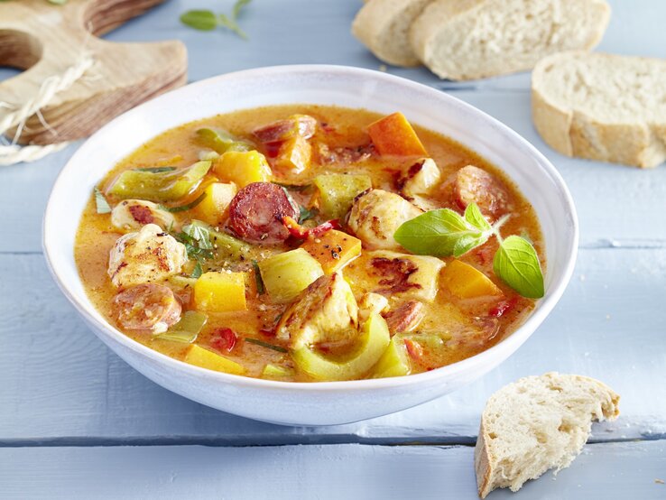 Hähnchen-Kürbis-Gulasch in einem weißten tiefen Teller. Daneben liegt Brot und ein Holzbrett. 