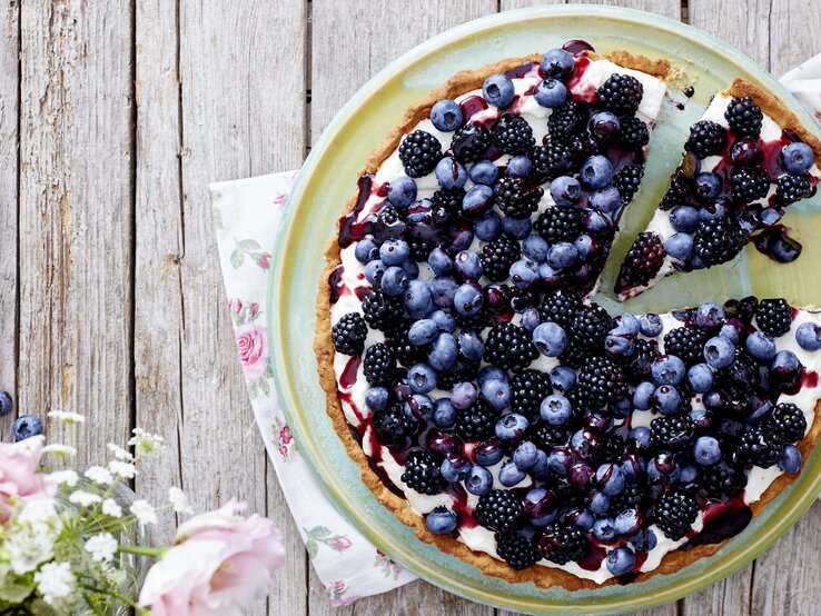 Eine Beeren-Tarte auf einem hellgrünen Teller. Unter dem Teller liegen Servietten mit Blumenmuster. Der Teller steht auf einem Holztisch. Am Bildrand befinden sich Blumen.