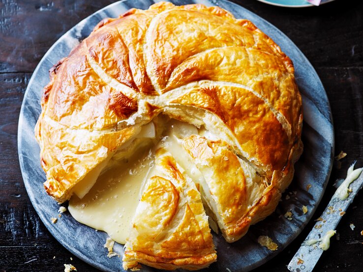 Ein großer Blätterteig-Pie mit Kartoffeln und Camembert auf einer blau-weißen, runden Scheibe, angeschnitten, sodass der geschmolzene Camembert hinausläuft, ein Stück fehlt, daneben ein Messer, von oben. 