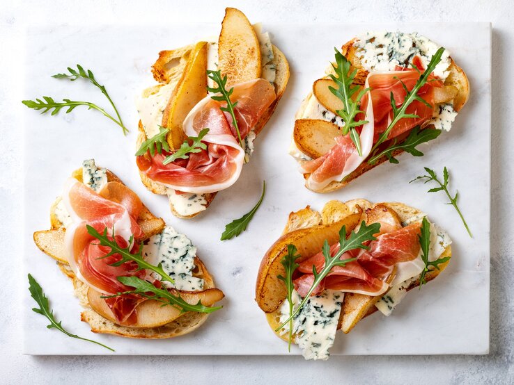 Crostini mit Birne, Schinken und Gorgonzola auf einer grauen Keramik-Platte. 