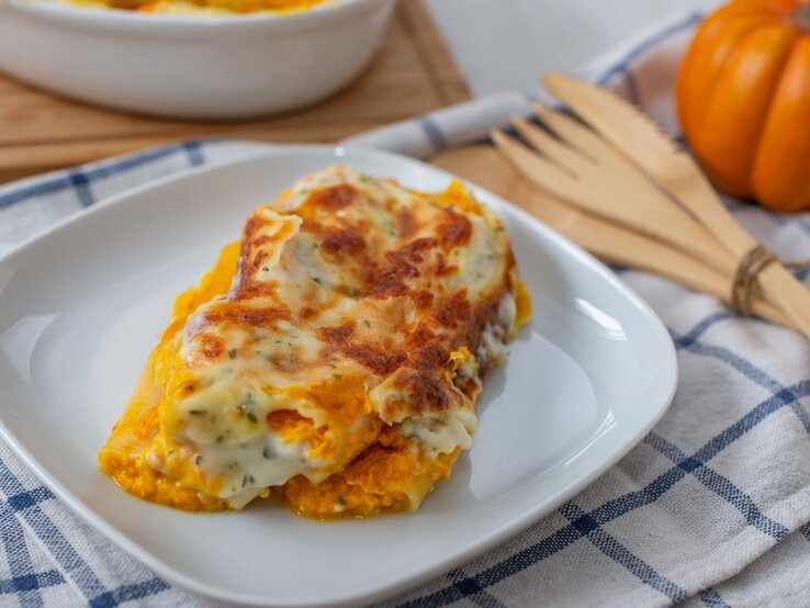 Kürbis-Cannelloni mit Kräuter-Bechamel und Mozzarella überbacken auf weißem Teller und blau-weißem Tuch aus der Draufsicht. Holzbesteck rechts mit Schnur zusammengebunden. Daneben kleiner Kürbis leicht verschwommen. Links oben angedeutete weiße Auflaufform auf Holzbrett.