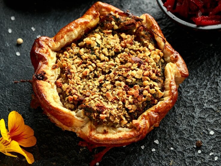 Eine Kürbis-Tarte mit Rote-Bete und Spinat garniert mit Walnüssen, Kürbiskernen und Fetakäse, gold-braun gebacken, neben einer Schüssel Rote-Bete und einer gelben Blume, vor dunklem Hintergrund.