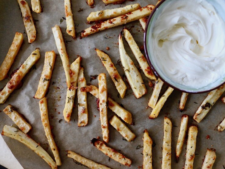 Auf einem Blech liegen Low-Carb-Pommes aus Pastinake. Daneben ist ein weißer Dip in einer Schüssel.