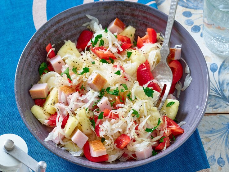 Sauerkrautsalat mit Kasseler in einer Schüssel auf einem blauen Untersetzer. Darunter eine Tischdecke mit blauen Blumen.