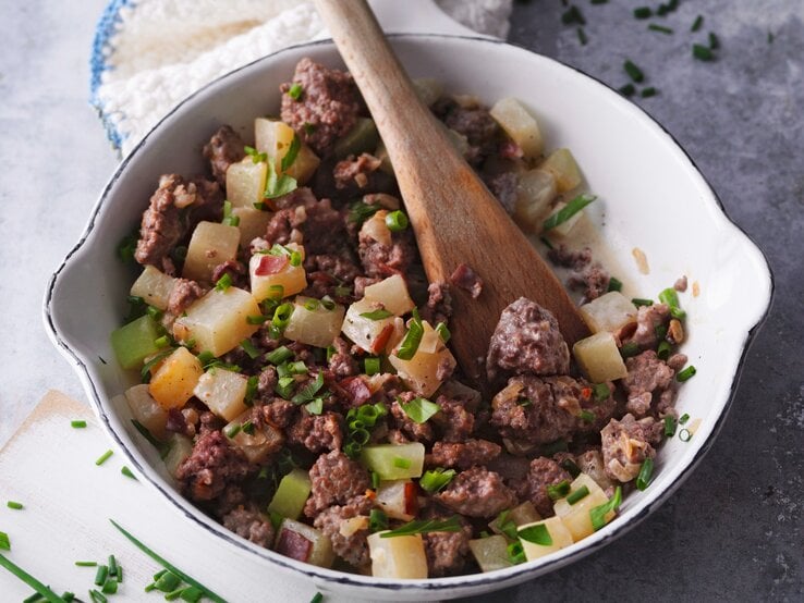 Draufsicht: Eine  weiße Hackfleischpfanne mit Kohlrabi. In dem Essen steckt ein großer Löffel aus Holz.