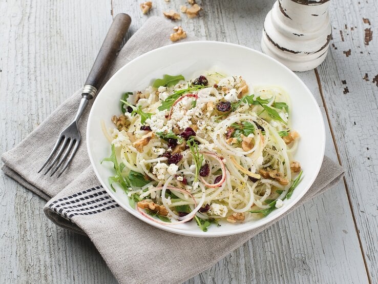 Draufsicht: Ein runder Teller mit einem Kohlrabi-Apfel-Salat mit Käse und Walnüssen auf einem hellen Untergrund aus Holz.