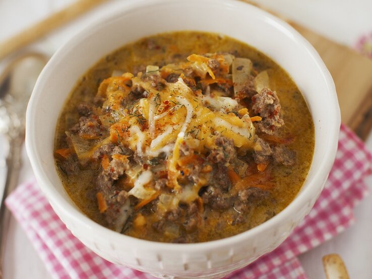 Eine weiße Schüssel mit herzhafter, cremiger Suppe aus Hackfleisch und Gemüse, garniert mit geschmolzenem Käse auf kariertem Tuch.