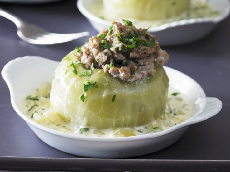 Gefüllter Kohlrabi mit Hackfleisch in einem tiefen weißem Teller auf grauem Untergrund. Im Hintergrund ist unscharf eine zweite Portion im Teller zu sehen.