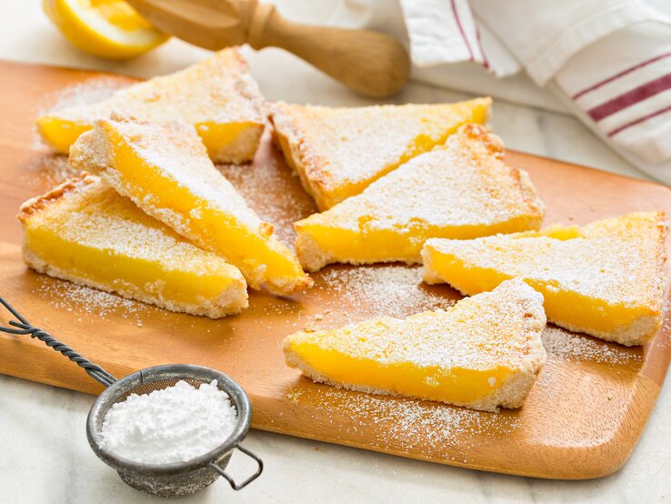 Zitronenkuchenstücke auf einem Holzbrett mit Puderzucker bestäubt, daneben ein Sieb und eine angeschnittene Zitrone im Hintergrund.