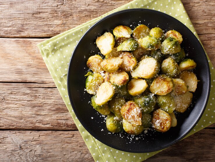 Draufsicht auf gebratenen Rosenkohl mit Parmesan auf schwarzem Teller. Dieser steht auf Holztisch.