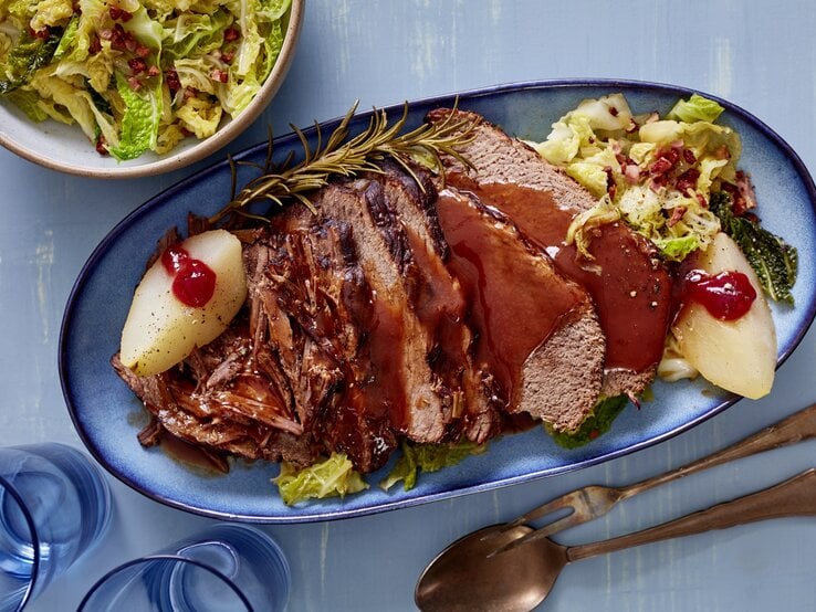 Rinderbraten mit Wirsing auf einem blauen Servierteller auf blauem Hintergrund. Unten auf dem Foto Besteck und Gläser, oben auf dem Foto eine Salatschüssel.