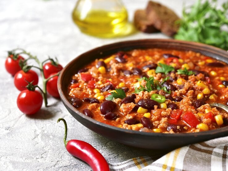 Dunkler Suppenteller gefüllt mit Chili con carne mit Mais und Kidneybohnen vor weißem Hintergrund. 