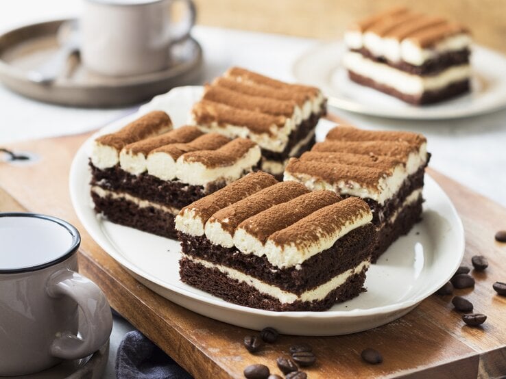 Ein weißer Teller mit drei Stücken der Tiramisu-Torte mit Mascarponecreme auf einem dunklen Holztisch.