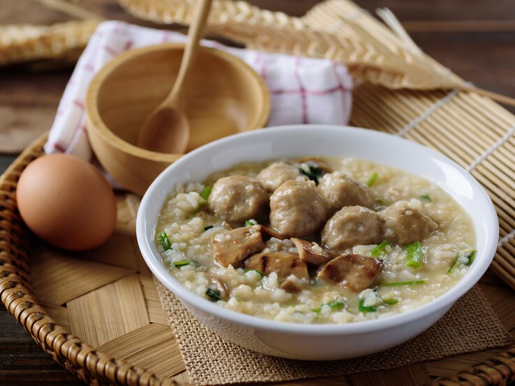 Reissuppe mit Hackbällchen & gebratenen Pilzen in weißer Schüssel auf hölzernem Untergrund.