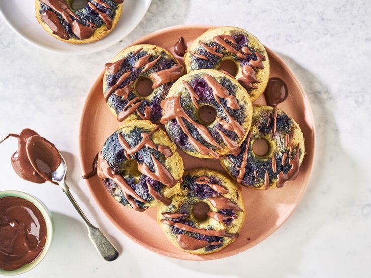 Draufsicht: Weißer Marmoruntergrund auf dem in der Bildmitte ein Tonteller mit sechs Donuts steht. Links daneben steht ein kleines Schüsselchen mit Mandel-Schokoladen-Topping und einem Löffel. In der oberen linken Bildecke steht ein weiterer, weißer Teller mit einem einzelnen Donut drauf.