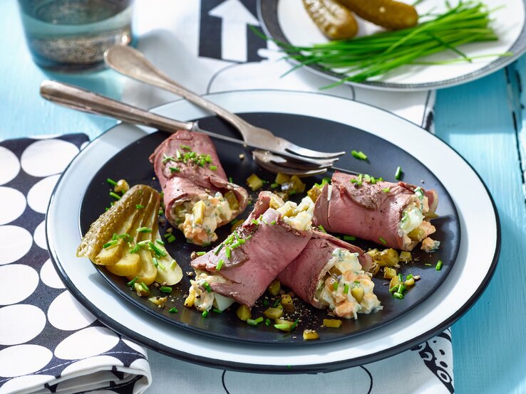 Roastbeef mit Eiersalat auf einem Teller auf einem schwarz-weiß-gemusterten Tischtuch. Hinten auf den Foto ein Teller mit Essiggurken und Schnittlauch. | © Matthias Liebich