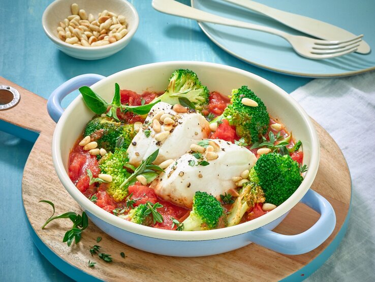 Kabeljaufilet mit Brokkoli und Pinienkernen in einem weiß-blauen Pfännchen auf einem hölzernem Brett. Der Hintergrund ist blau und hinten auf dem Foto Messer und Gabel.