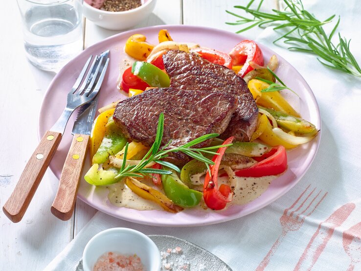 Rindersteak mit Paprikagemüse auf einem rosa Teller, links das Besteck. Der Hintergrund ist weiß.