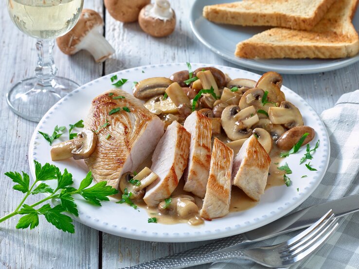 Putenschnitzel mit Champignons auf einem weißen Teller. Rechts Besteck und links ein Weinglas. Der Hintergrund ist hell und Champignons darauf verteilt.