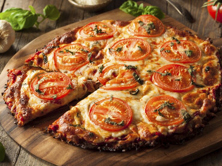 Nahaufnahme der Blumenkohl-Pizza auf einem rustikalen Holztisch mit Tomaten und Basilikum.