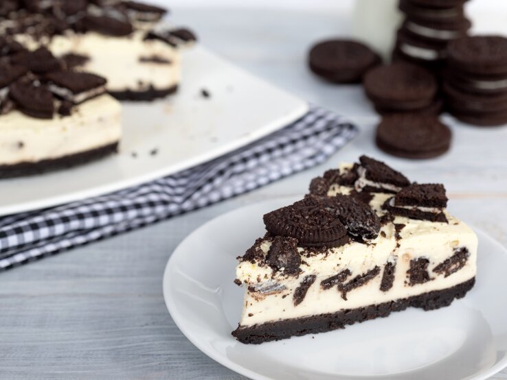 Ein Stück cremiger Oreo-Kuchen auf weißem Teller, daneben der restliche Kuchen auf kariertem Tuch und gestapelte Oreos im Hintergrund.