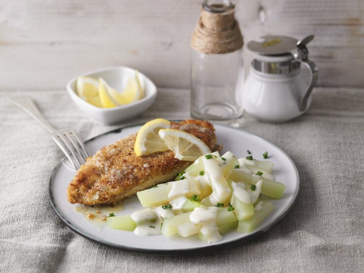 Ein weißer Teller mit dem Mandel-Schnitzel und Kohlrabi-Rahm auf einem hellen Tisch mit Zitronenecken garniert.