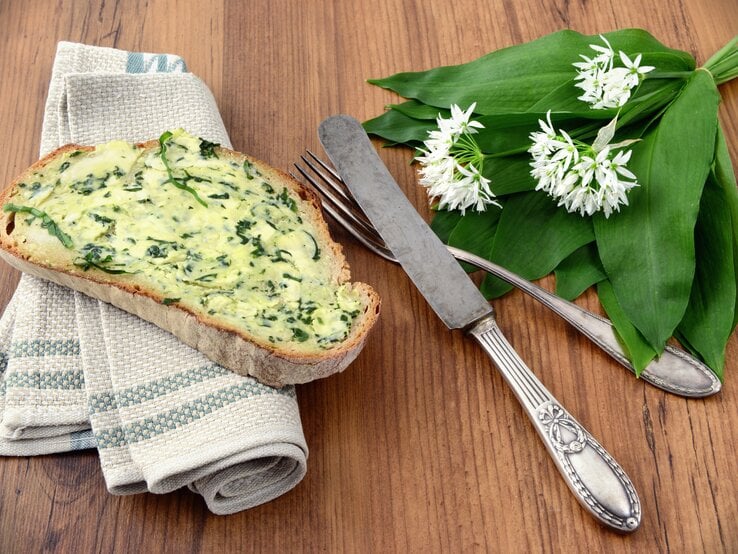 Brotscheibe beschmiert mit Bärlauchbutter auf Holztisch, daneben Besteck.