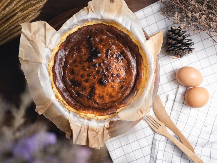 Frisch gebackener, leicht verbrannter Käsekuchen auf Backpapier, umgeben von Eiern und Naturdeko auf karierter Stoffserviette.