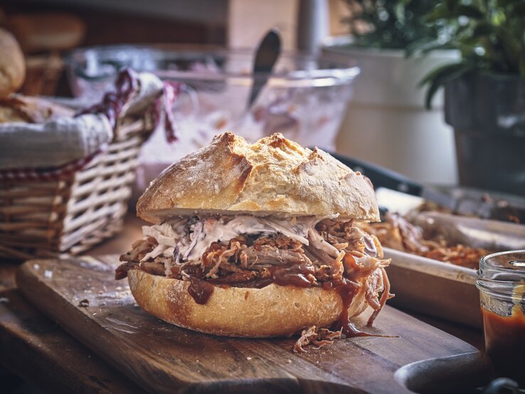 Saftiges Pulled Pork Sandwich mit Krautsalat auf einem rustikalen Holzbrett, im Hintergrund ein geflochtener Brotkorb.