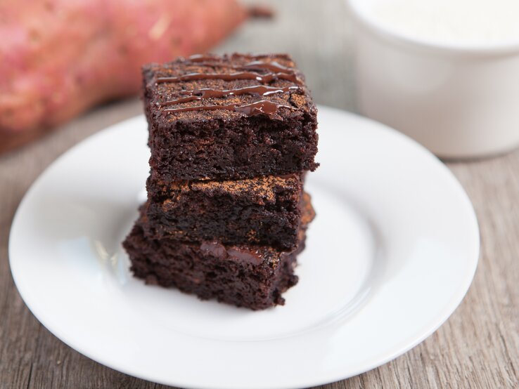 Drei Süßkartoffel-Brownies auf einem weißen Teller in der Nahaufnahme. Alles auf grauer Tischfläche und eine Süßkartoffel unscharf im Hintergrund erkennbar.