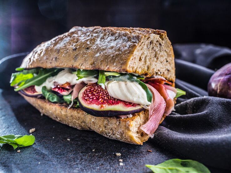 Frontalsicht: Schinken-Mozzarella-Sandwich mit Salatblättern liegt auf einem dunklen Untergrung. Daneben ist ein dunkles Tuch.