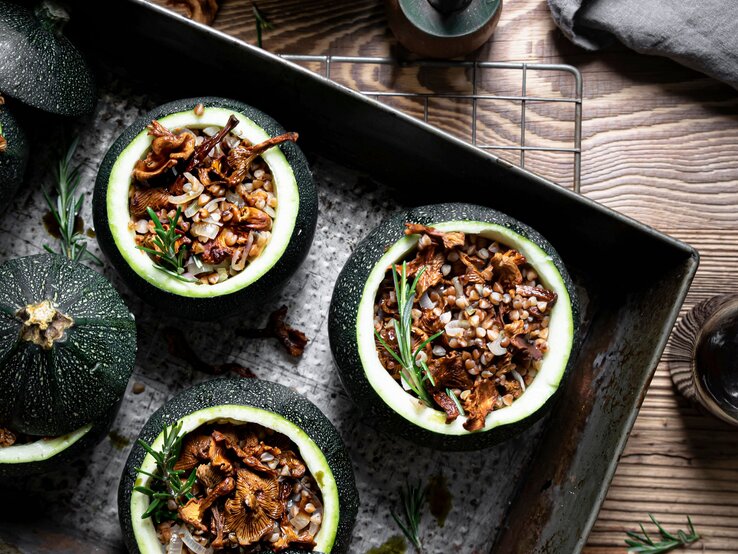 Draufsicht: Vier runde gefüllte Zucchini mit Pfifferlingen und Buchweizen auf einem dunklem Tablett, dass auf einem Holztisch liegt. Daneben steht ein Pfeffermühle sowie ein Schale mit der Zucchini-Füllung.