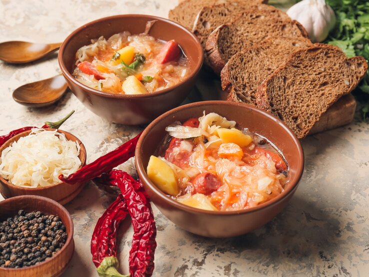 Draufsicht: Auf einem Untergrund aus Mamor stehen zwei Schüsseln mit einer Sauerkraut-Kassler-Suppe. Drum herum liegen Brotscheiben, Holzlöffel, rote Chilischoten, eine Schale Pfefferkörner und frisches Sauerkraut.