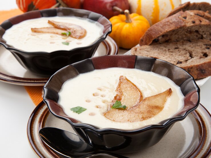 Zwei dunkle Schüsseln mit Pastinaken-Suppe, dekoriert mit Birnenscheiben, Ahornsirup und Petersilie, daneben Brotscheiben und Kürbisse.